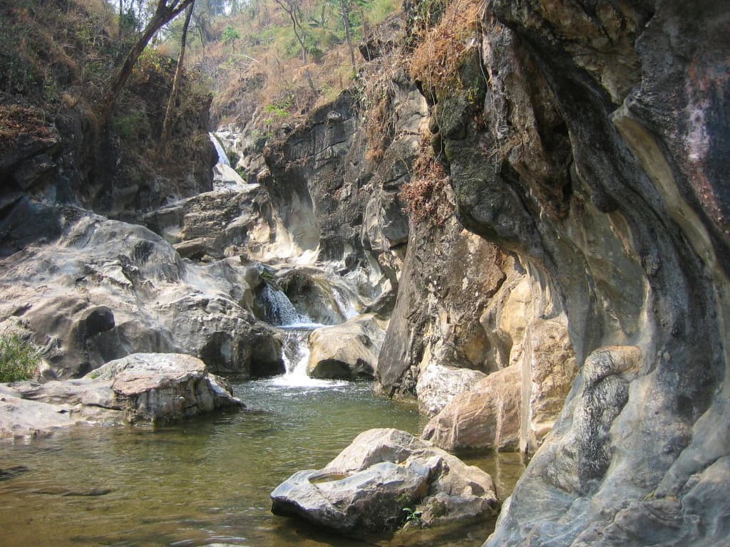 Rocky waterfall with small stream flowing over rocks into a pool, surrounded by vegetation and cliffs
