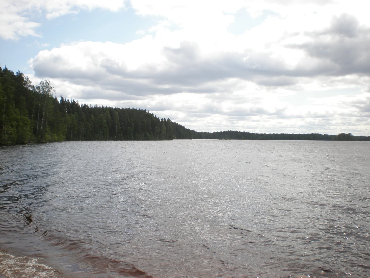Calm lake with pebbly shore, forested shoreline, and partly cloudy sky