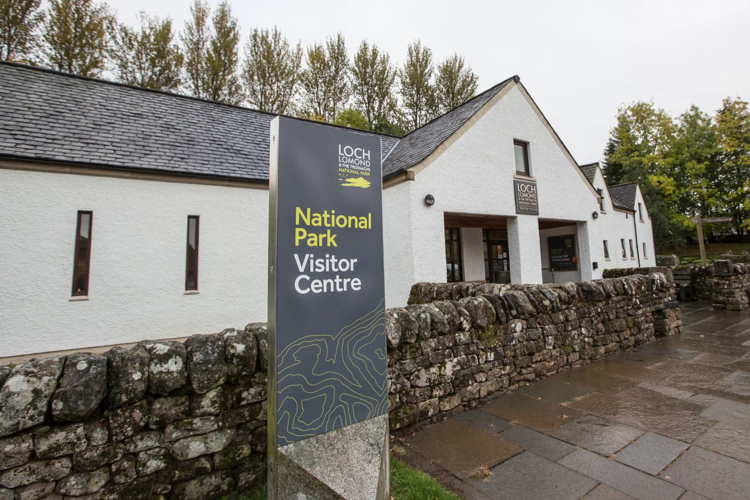 Sign reading 'National Park Visitor Centre' with 'Loch Lomond' logo, white building with stone wall, and trees in background