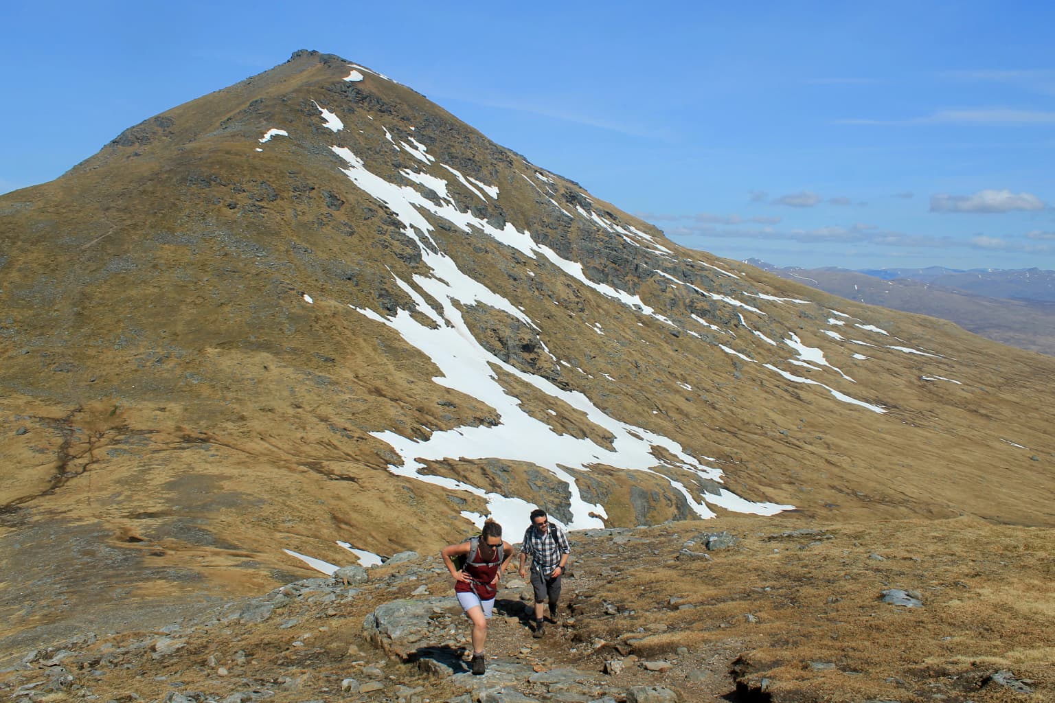 Two hikers on a rocky mountain ridge with Ben More mountain featuring snow patches in the background under a clear blue sky