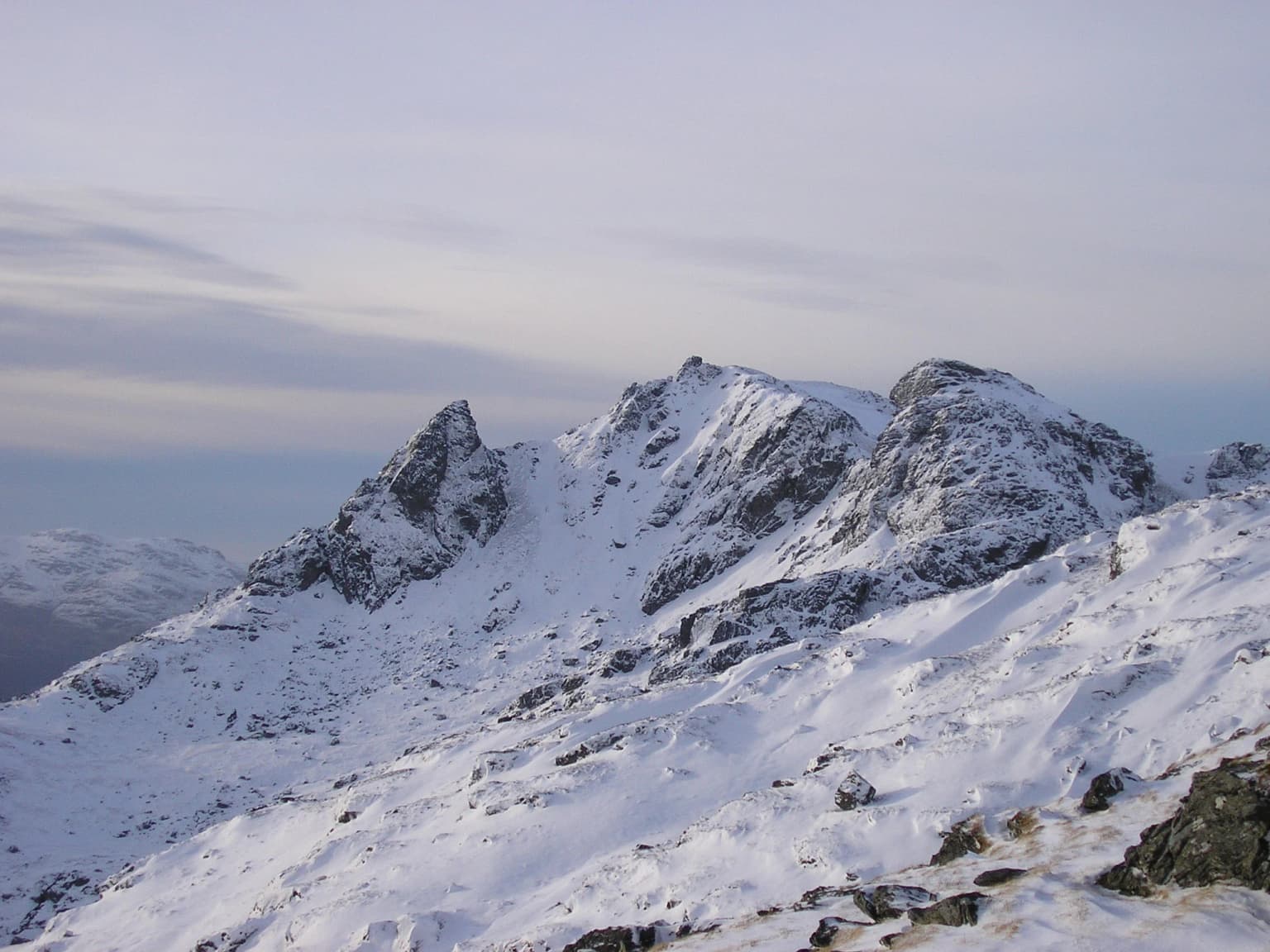 Snow-covered rocky mountain peaks under a pale sky, with snow-covered slopes and exposed rock faces