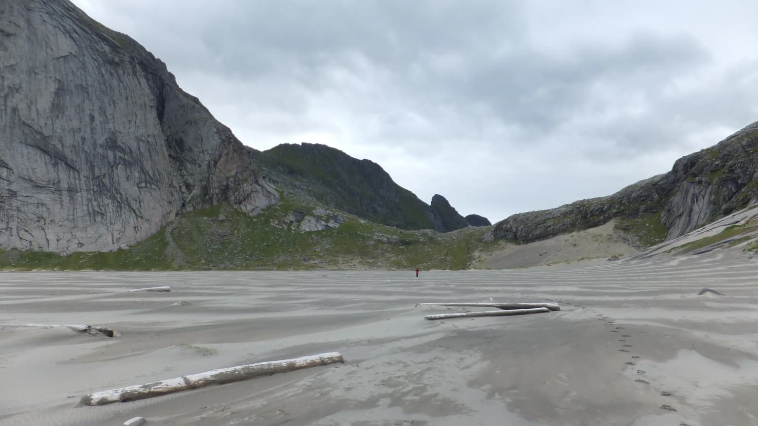 Bunes Beach in Lofotodden National Park, Norway, featuring sandy shore with driftwood, distant mountains, and a single figure under overcast sky