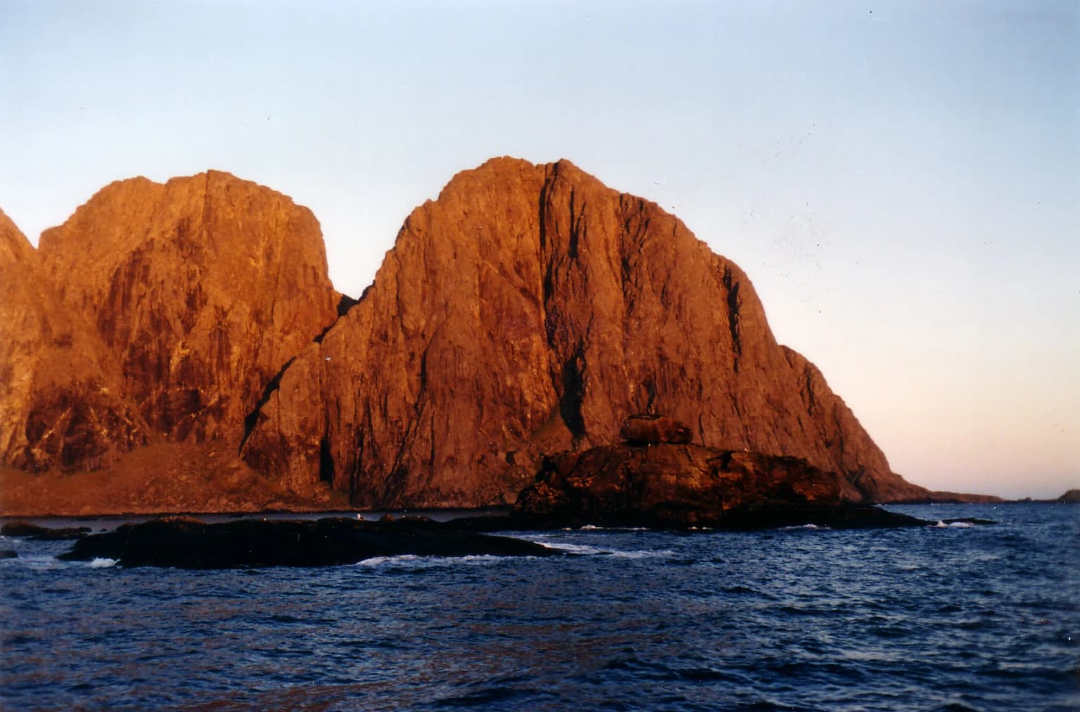 Large rocky mountain cliffs rising from the sea, illuminated by golden light during what appears to be sunrise or sunset