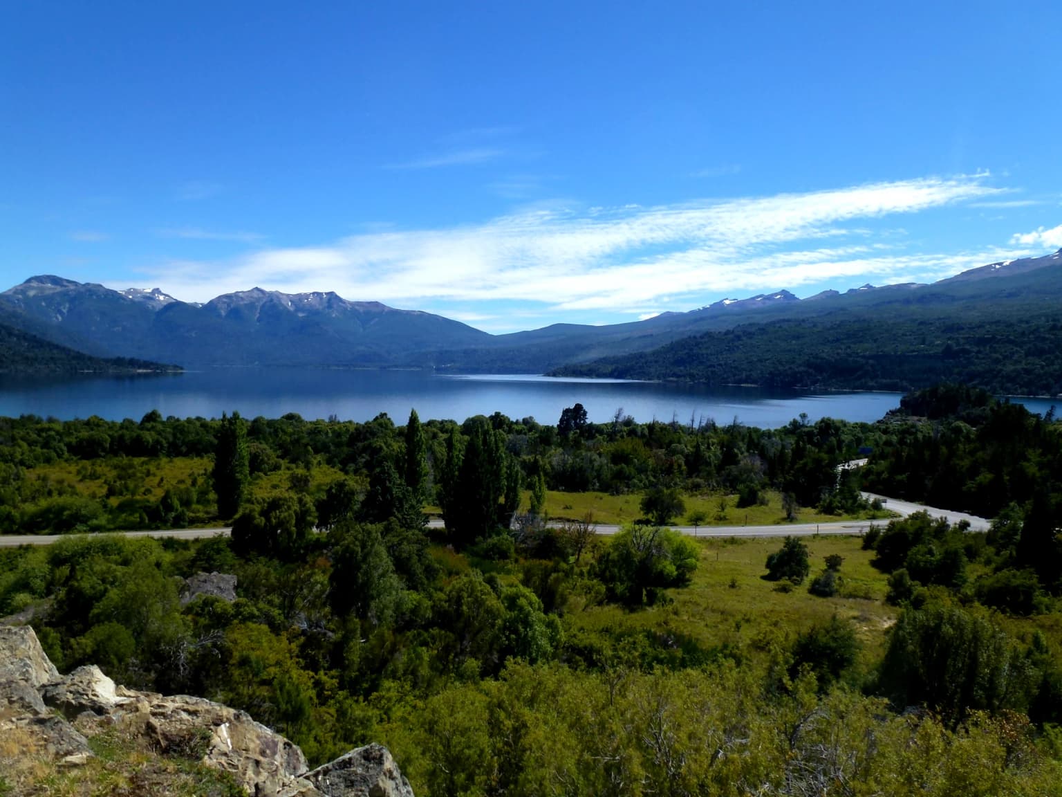 Lago rodeado de densos bosques verdes y montañas bajo un cielo azul despejado, con un camino visible a media distancia