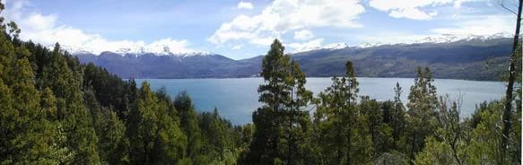 Vista panorámica del lago Futalaufquen con exuberantes bosques verdes en primer plano y montañas distantes bajo un cielo parcialmente nublado.