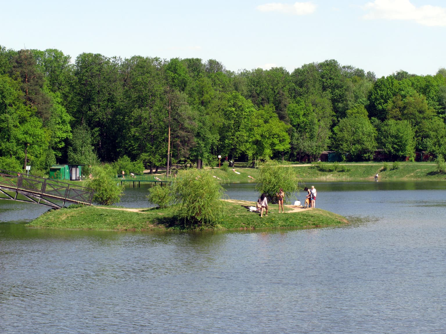 A pond with a small grassy island in the center where several people are standing, a wooden bridge on the left side, and dense green forest in the background under a partly cloudy sky.