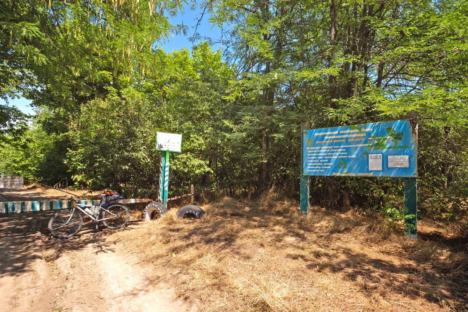 Dirt path leading to blue informational sign and smaller signpost with bicycle parked nearby, surrounded by trees