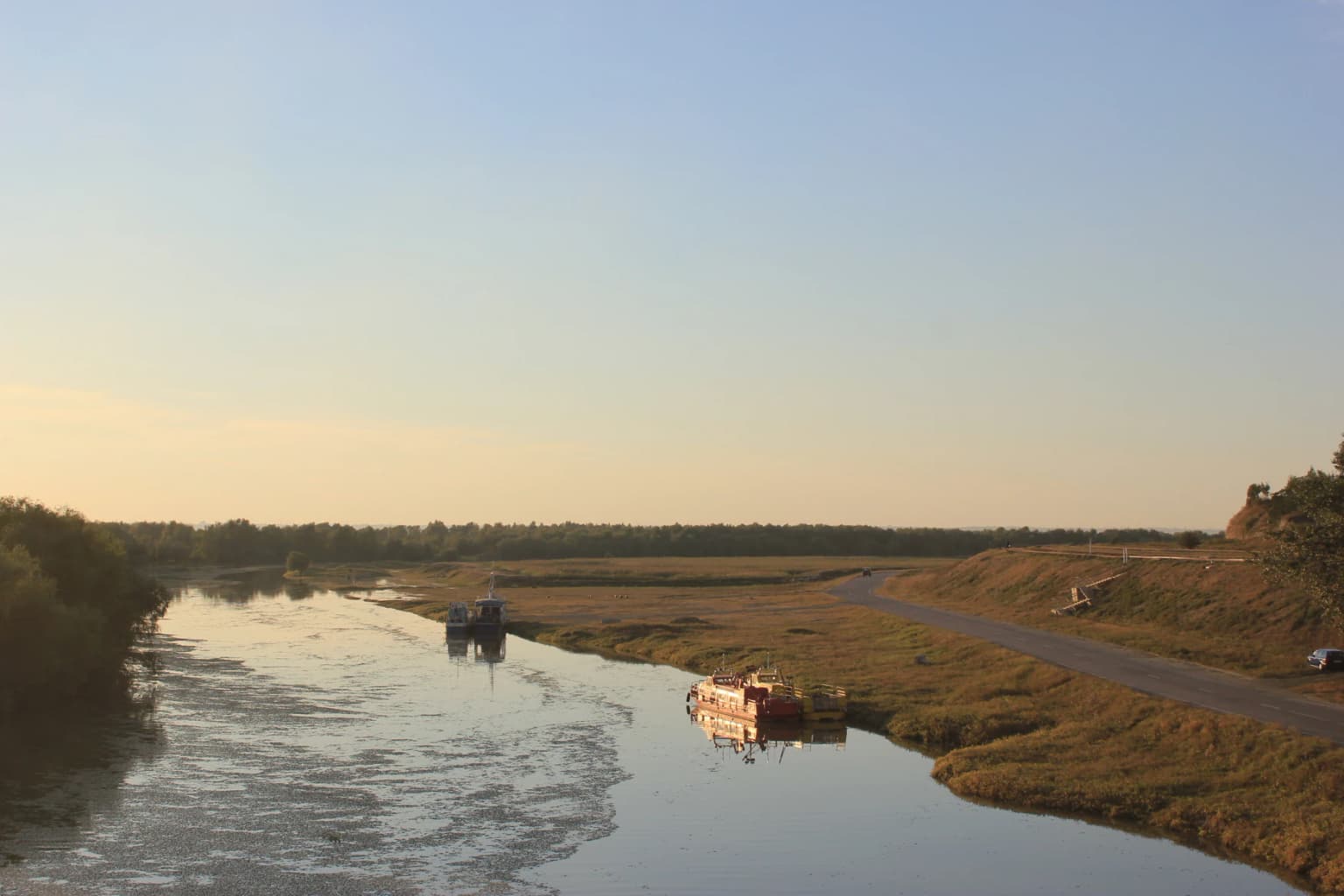 Wide view of a river with boats on calm water, grassy riverbanks, and a road on the right side of the image