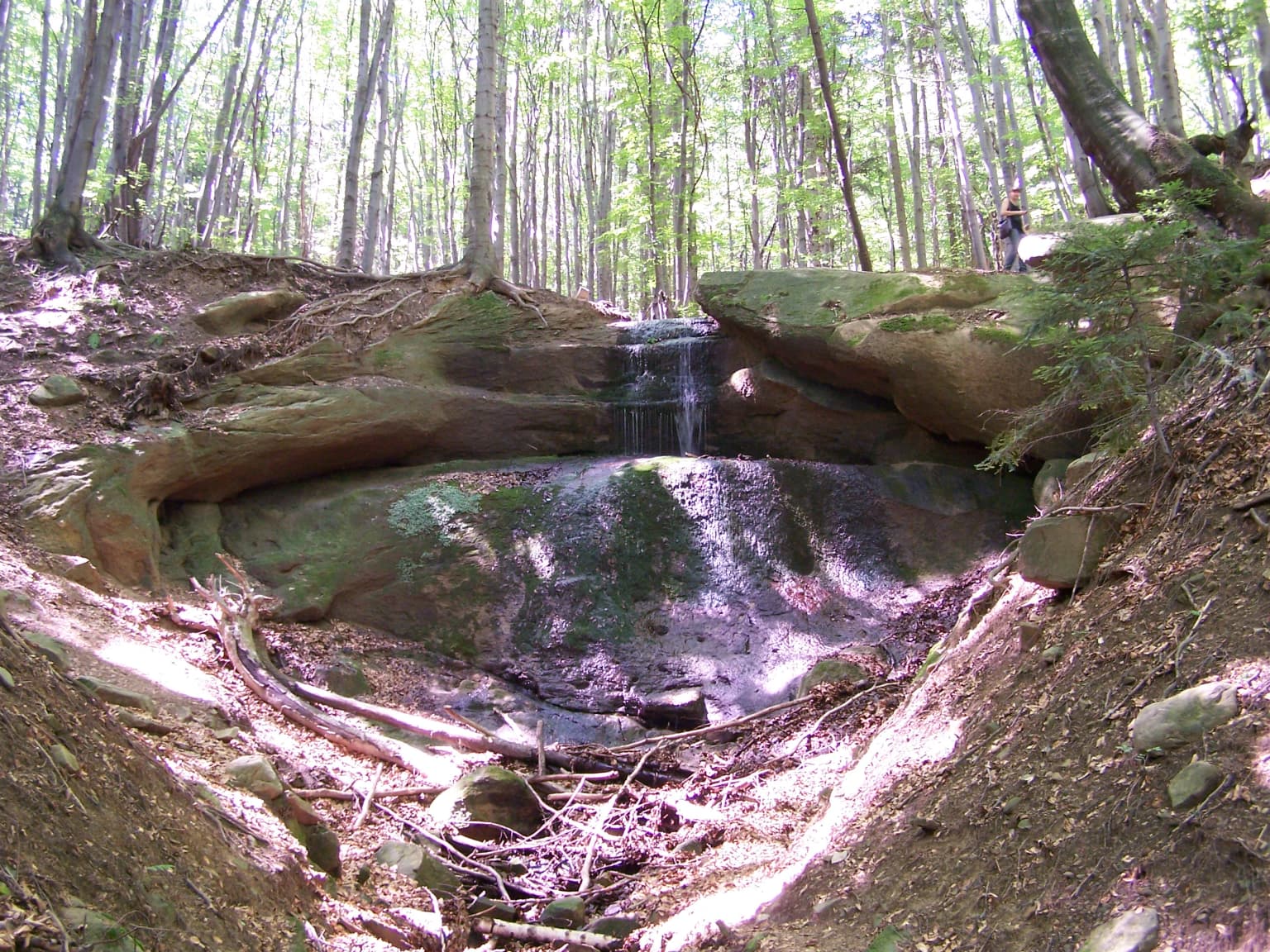 Waterfall cascading over rocks in a forested area with a person standing on a rock in the background