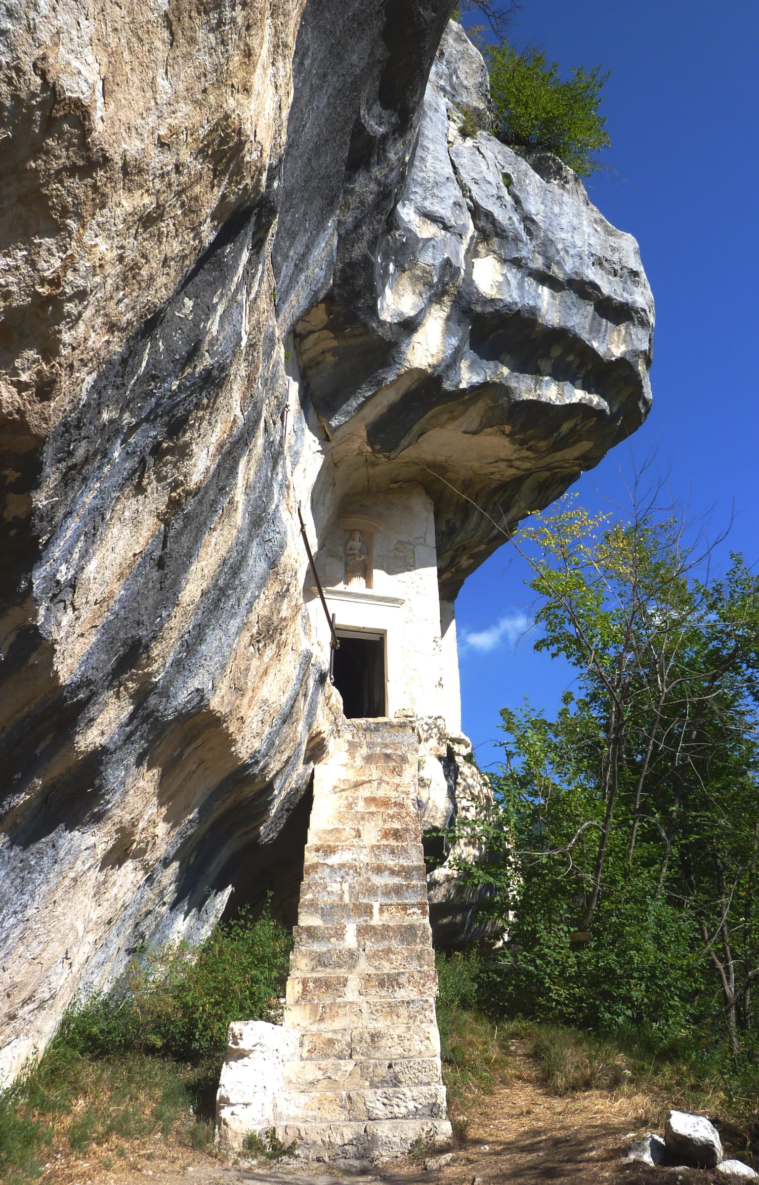 Stone hermitage built into a rock face with a stone staircase leading to an entrance, surrounded by greenery and a clear blue sky