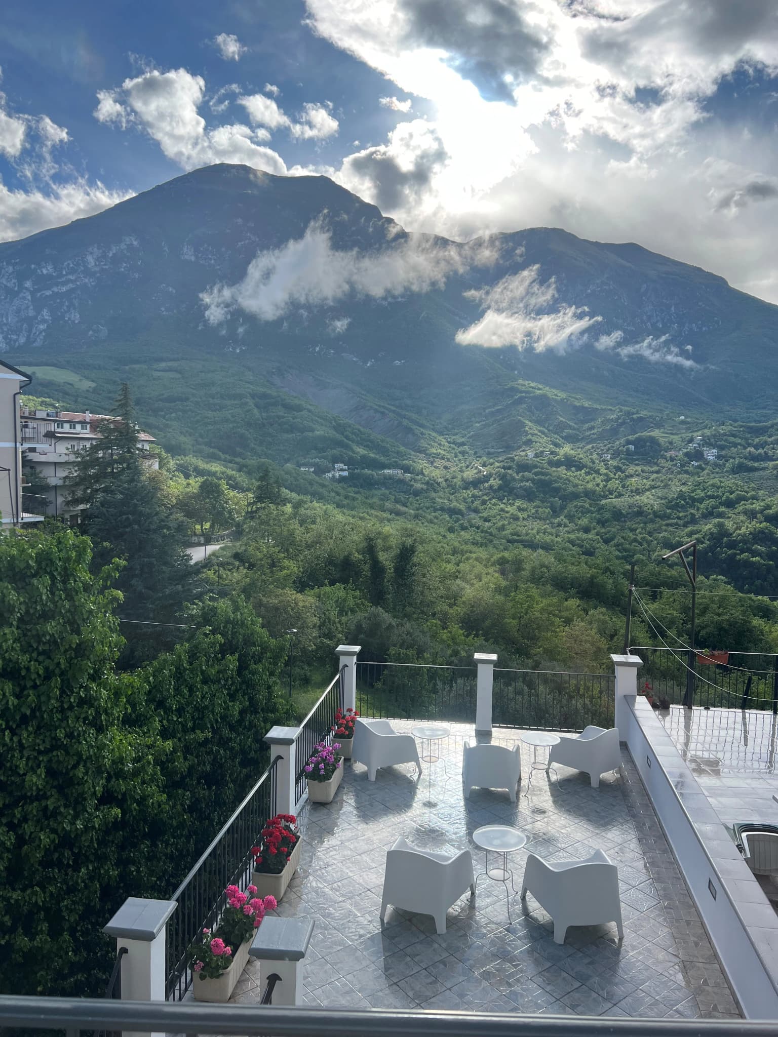 White patio with chairs and planters holding pink flowers, overlooking green mountains under a partly cloudy sky