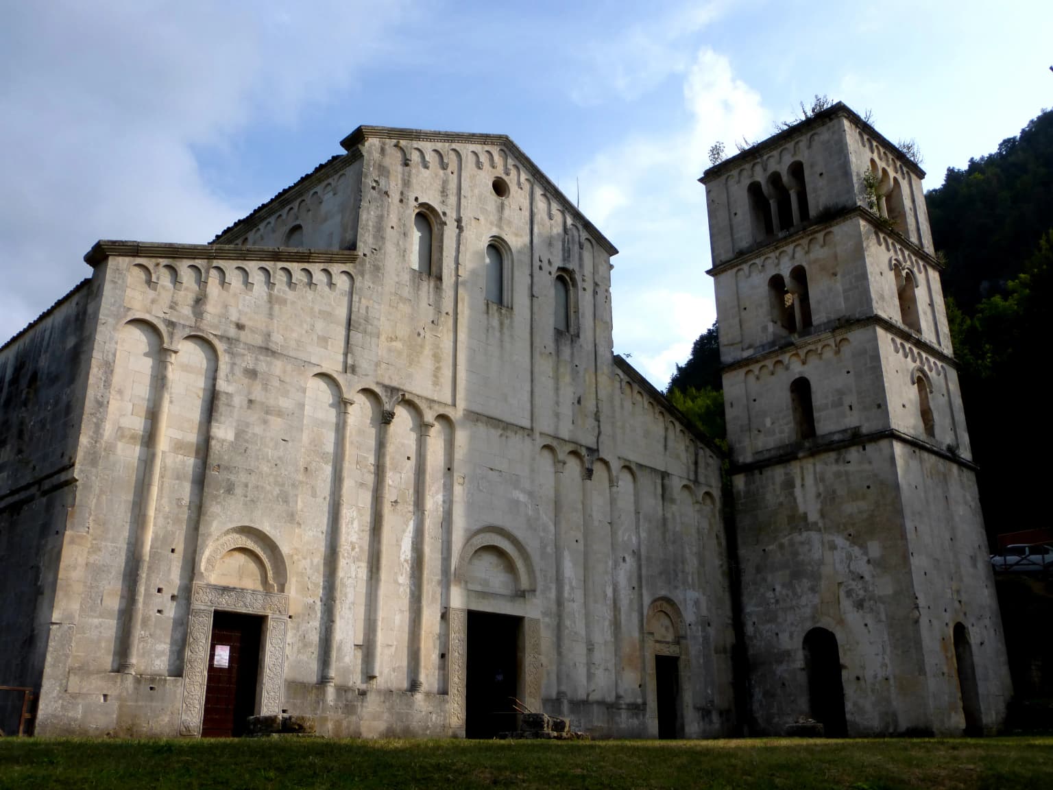 Stone abbey building with a tall tower, arched windows, and grassy field in front under a partly cloudy sky
