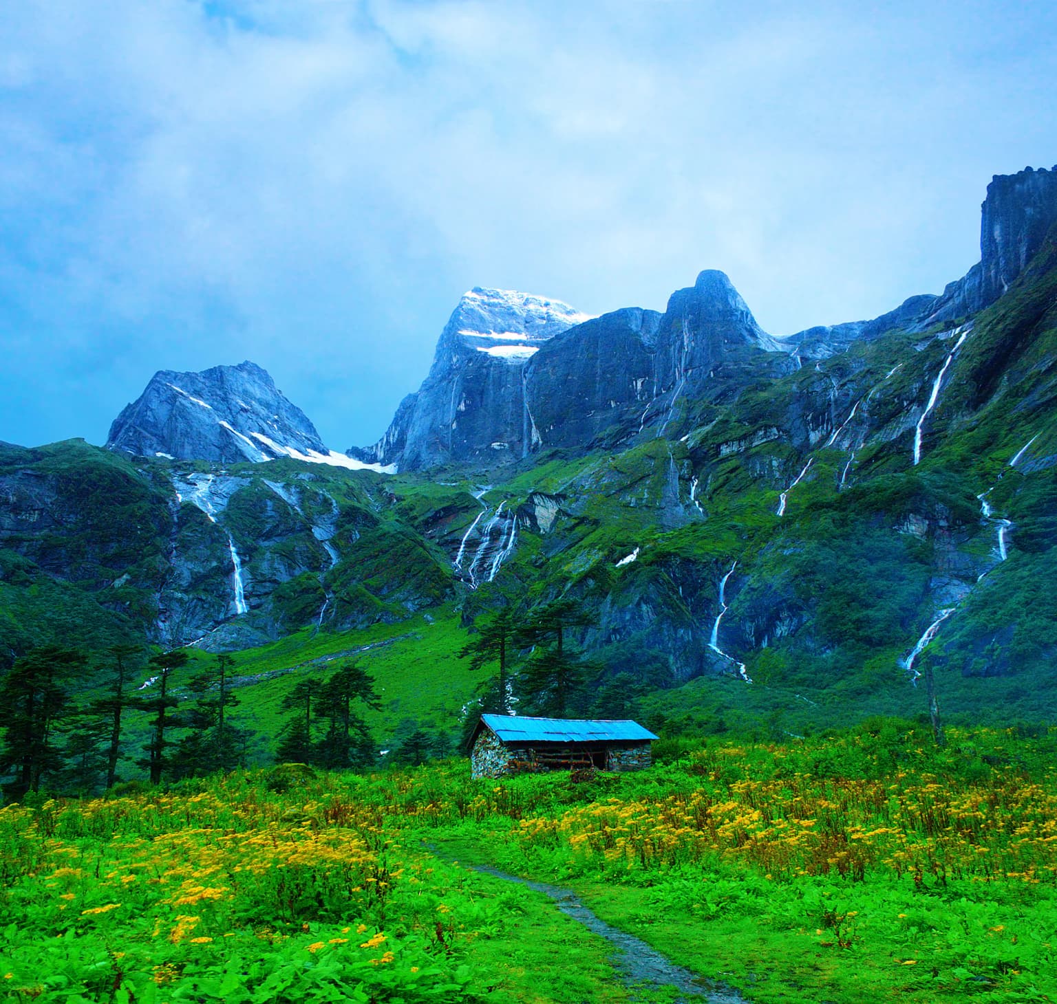 Barun Valley landscape featuring snow-capped mountains, multiple waterfalls, a small wooden hut with a blue roof, and a trail through a lush green meadow with yellow wildflowers