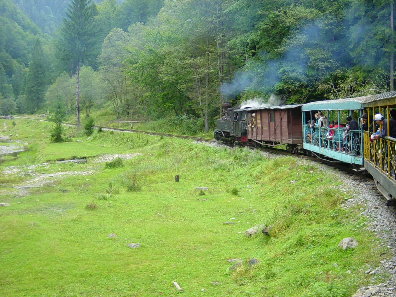 Steam train with passengers on a railway track through a lush green valley with trees and mountains