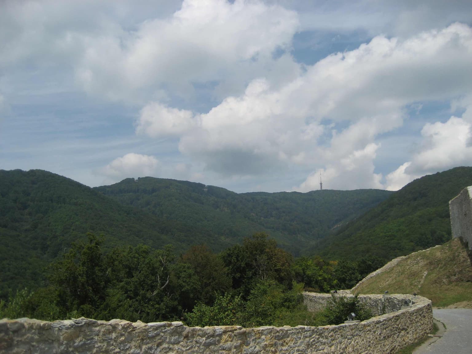 Stone wall in foreground, forested hills and mountains in midground, cloudy sky in background