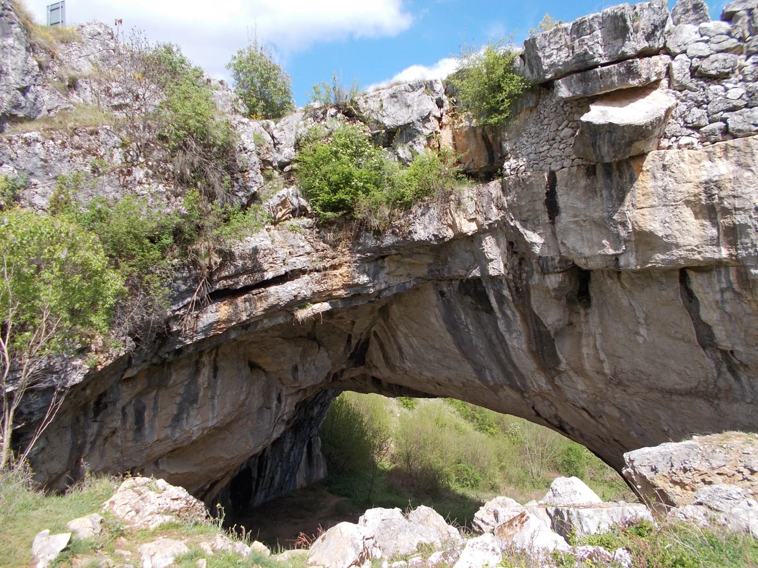 Natural stone arch bridge with vegetation on top and rocky terrain in the background under a blue sky