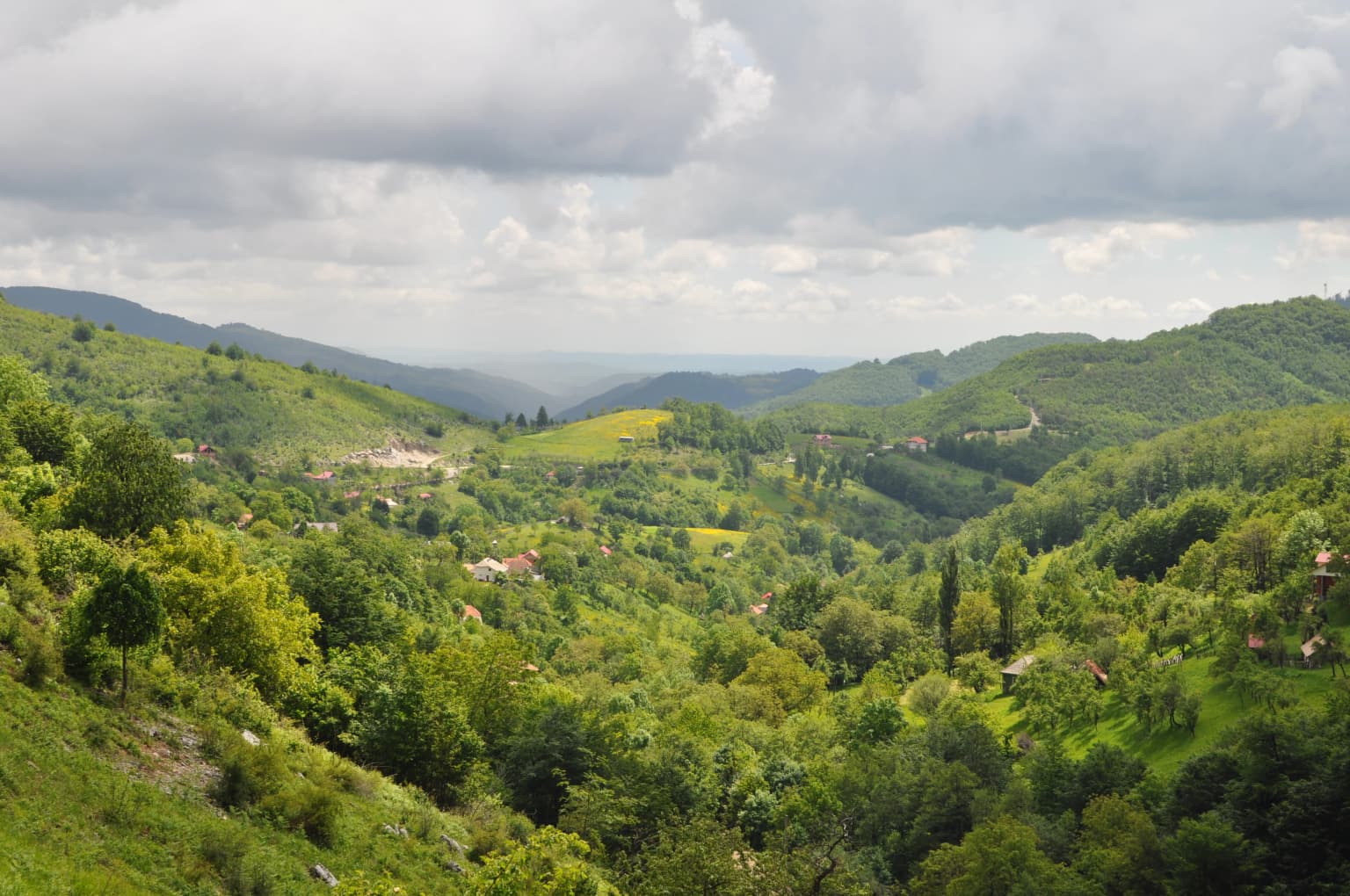 Wide landscape view of rolling green hills and valleys with scattered buildings under a partly cloudy sky