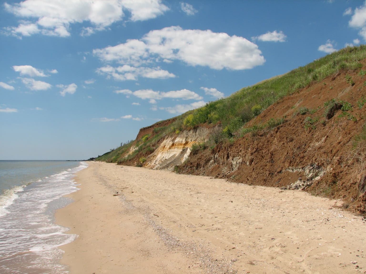 Sandy beach with shallow waves, coastal cliffs with green vegetation, and blue sky with scattered clouds.