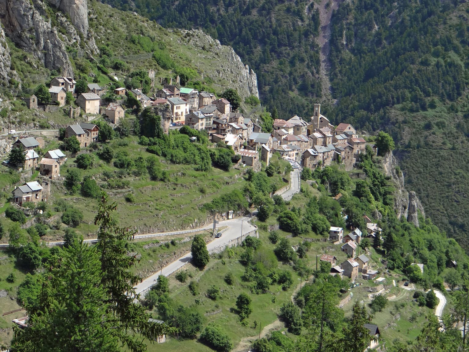 A village with stone houses on a hillside surrounded by green mountains and a winding road