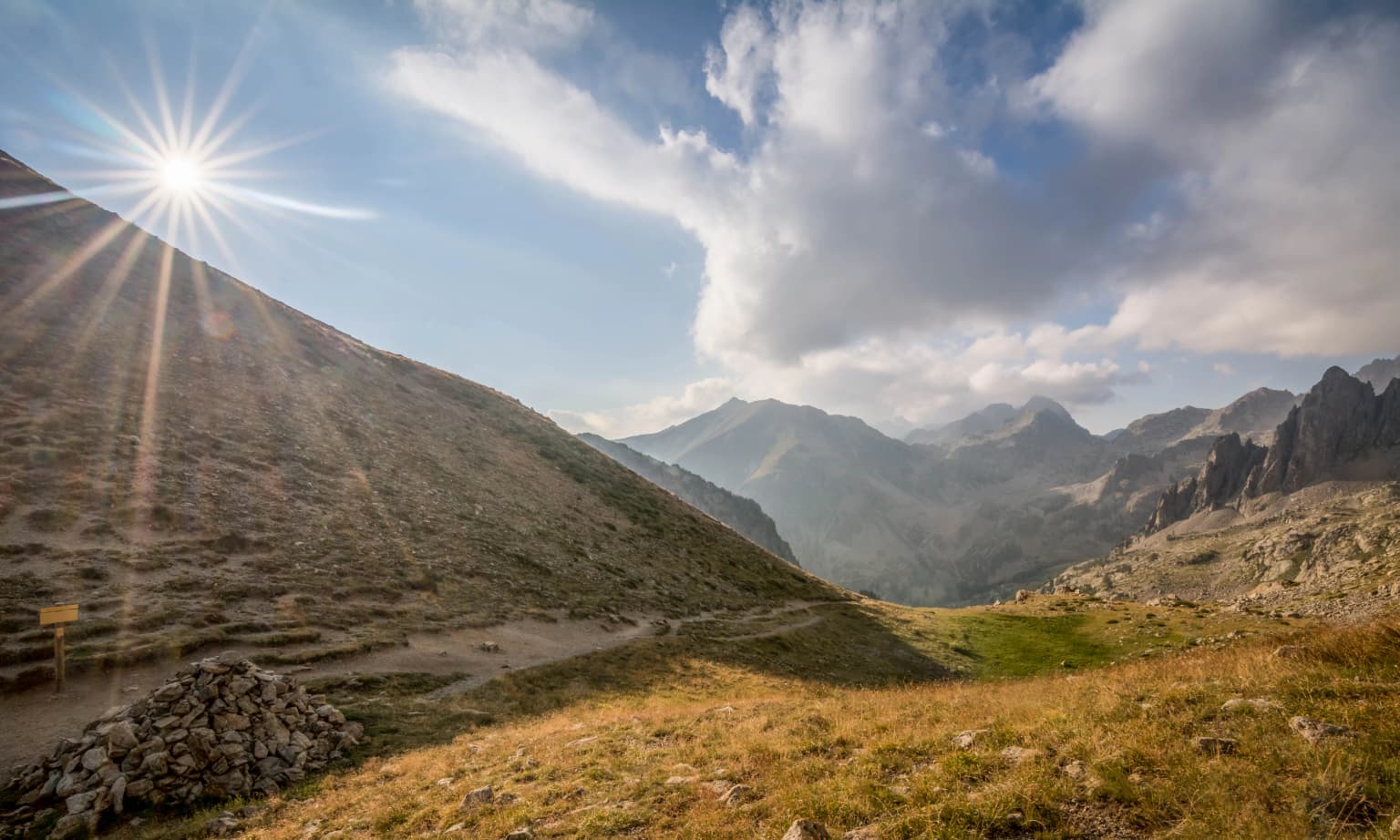 Sunrise over a mountain valley with rocky slopes and distant peaks under a partly cloudy sky