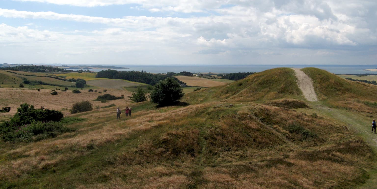Grassy hills with a dirt path, two people walking, scattered trees, and the sea under a partly cloudy sky