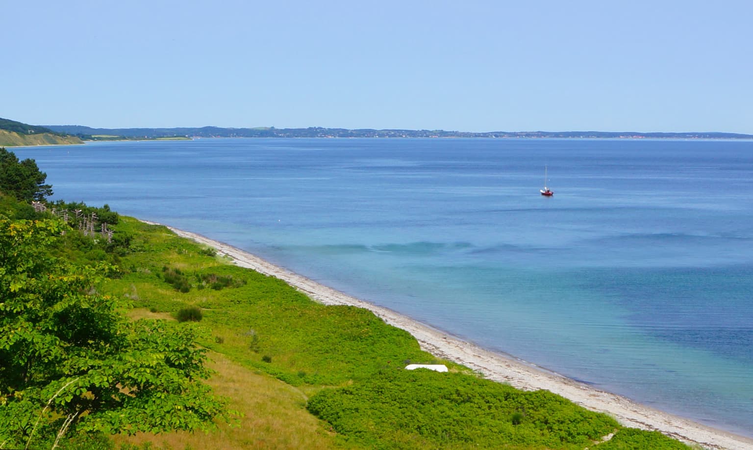 Coastal landscape featuring a sandy beach, calm blue water, green hills, and a small sailboat under a clear sky