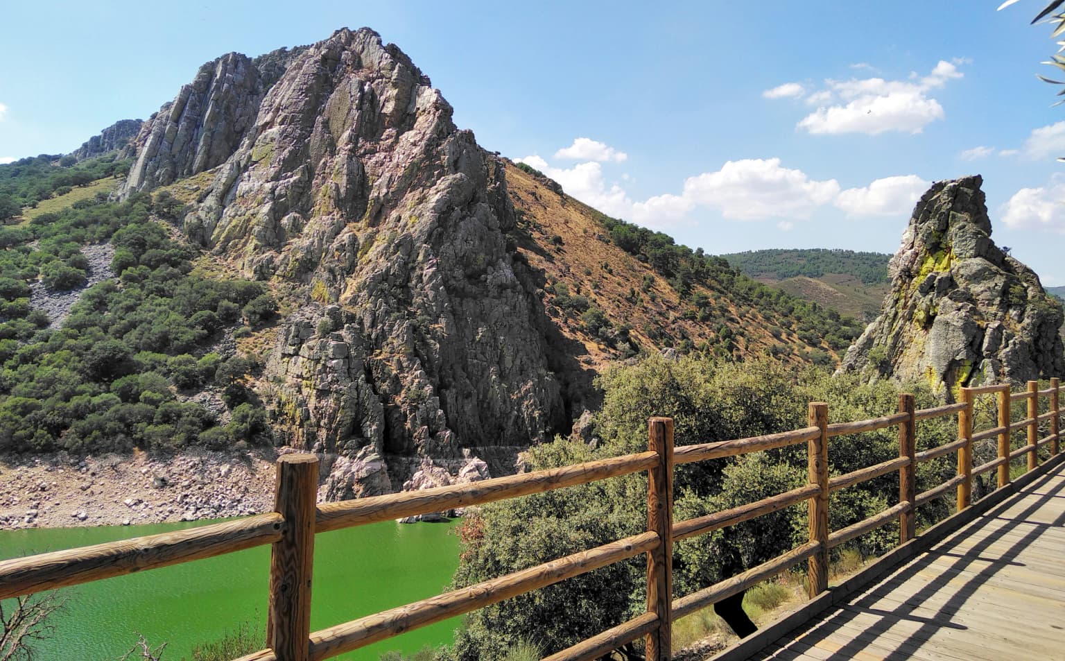Wooden railing of a viewpoint overlooking a green river with rocky mountains in the background under a partly cloudy sky