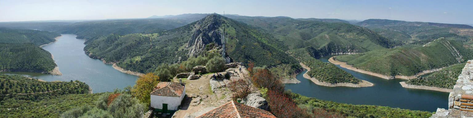 Panoramic view showing a river winding through mountainous terrain with scattered structures including Monfragüe Castle in the foreground