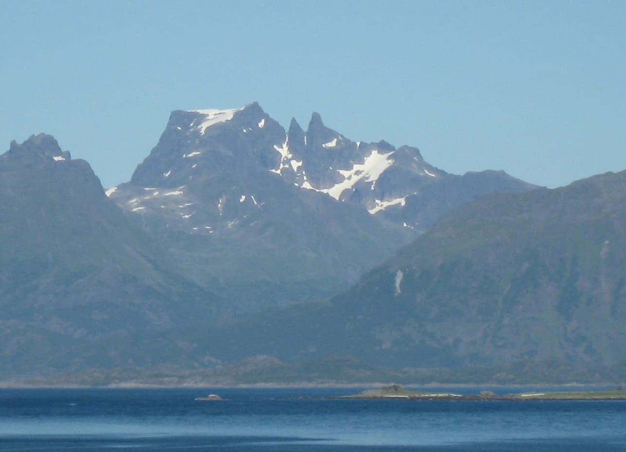 Snow-capped Møysalen Mountain rising above a calm body of water with surrounding hills under a clear blue sky