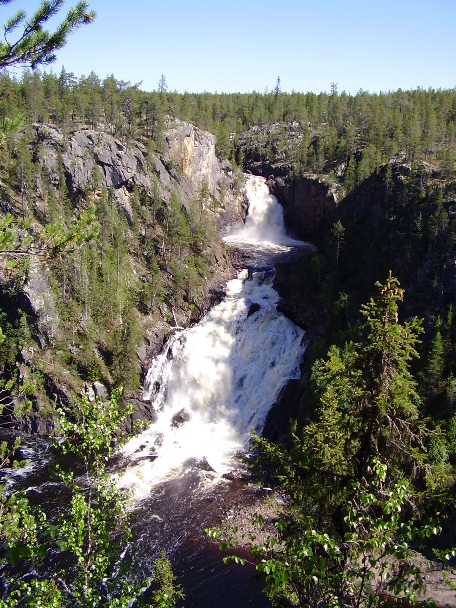 A waterfall cascading over rocks with dense forest in the background under a clear blue sky