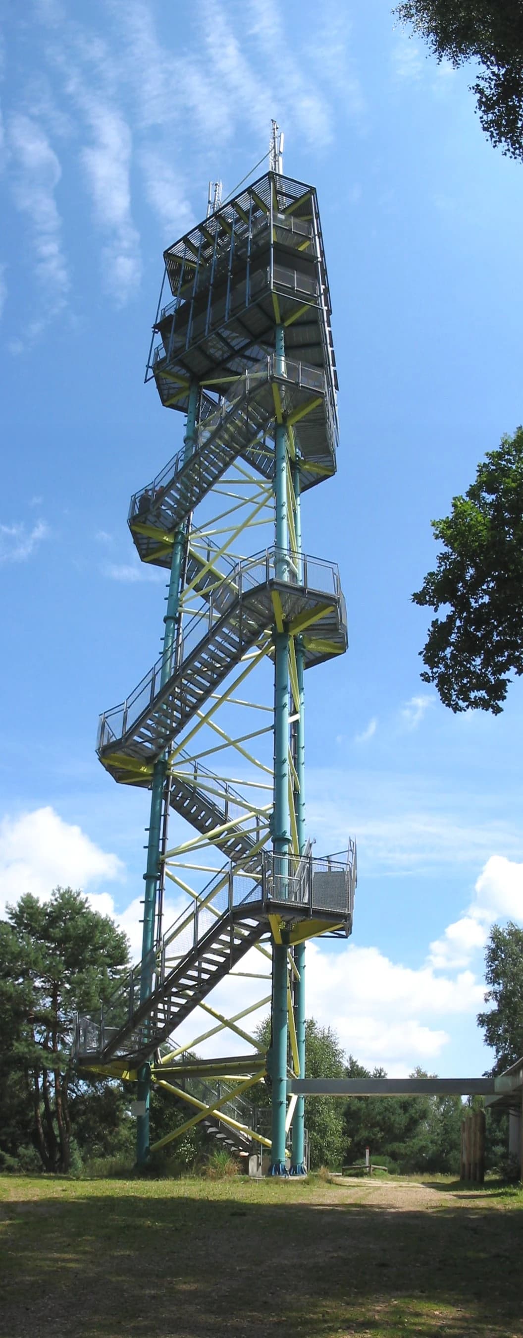A tall metal observation tower with spiral stairs and multiple viewing platforms surrounded by trees and grass under a clear blue sky.