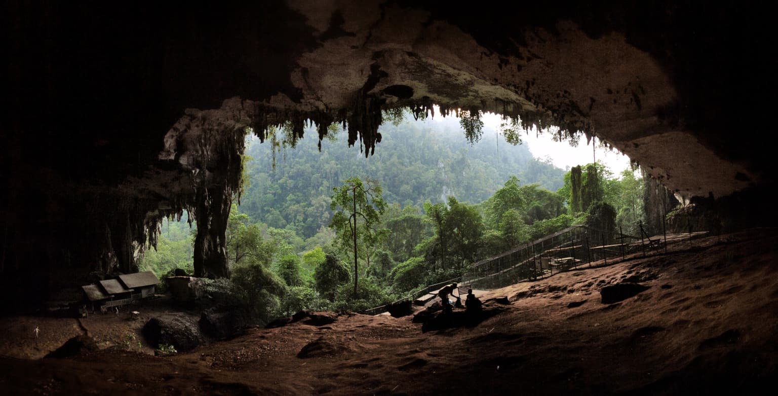 Rocky cave entrance with forest landscape visible beyond at sunset.