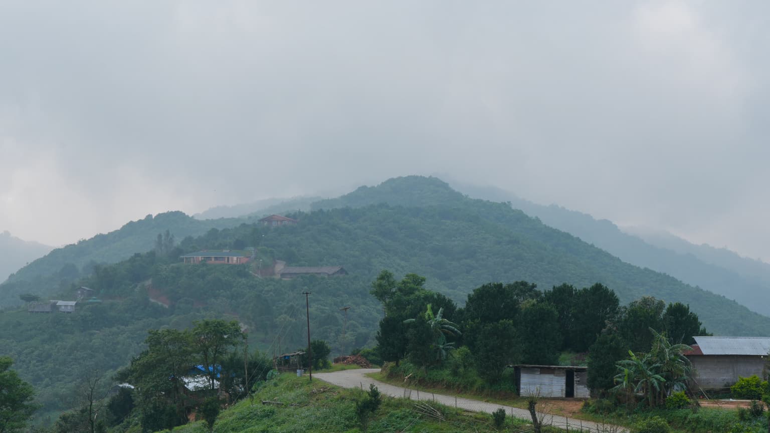 Loma montañosa verde y exuberante bajo un cielo nublado, con una carretera y pequeños edificios en primer plano