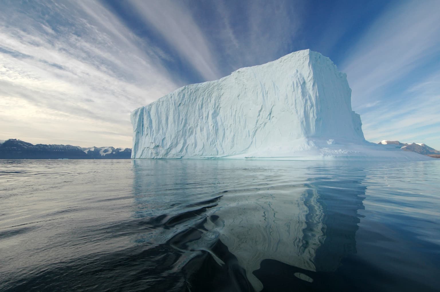 Large rectangular iceberg floating in calm water with reflection, under partly cloudy sky with distant landmasses visible