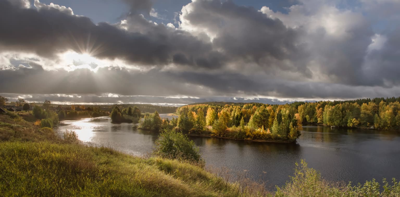 Panoramic view of a river with autumn-colored trees, grassy hills, and dramatic clouds with sunlight breaking through