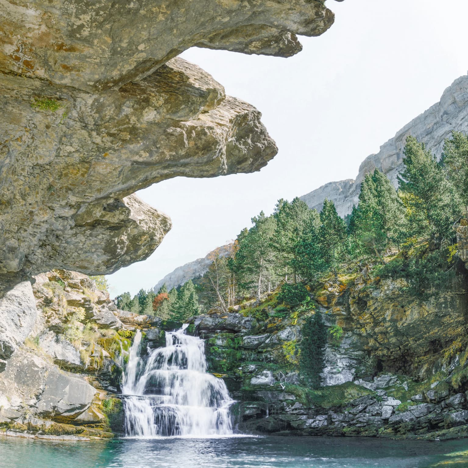 Waterfall flowing under rocky overhang with surrounding mountains and trees