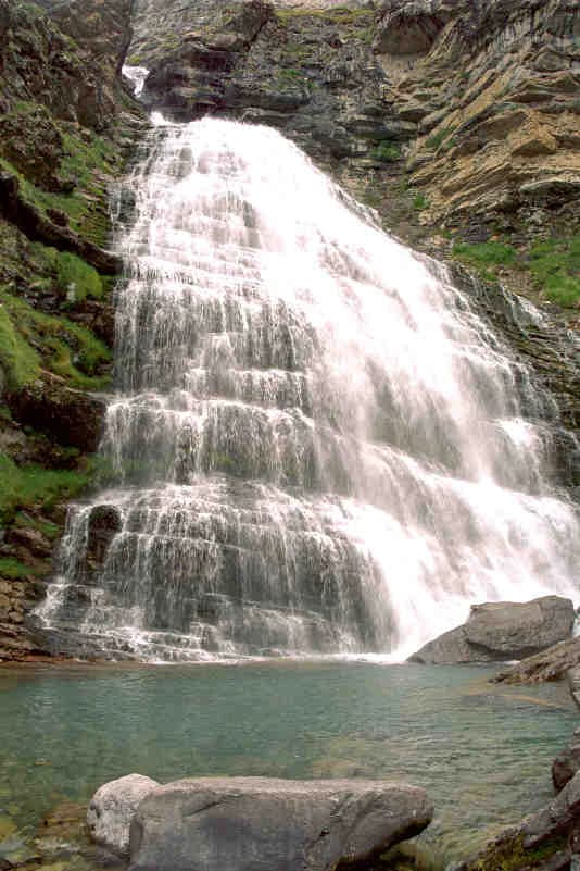 Waterfall cascading down rocky cliffs into a turquoise pool with green slopes and large boulders in the foreground