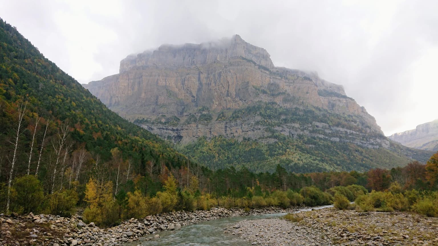 Mountainous landscape featuring Punta Tobacor peak with a river flowing through a forested valley under overcast sky