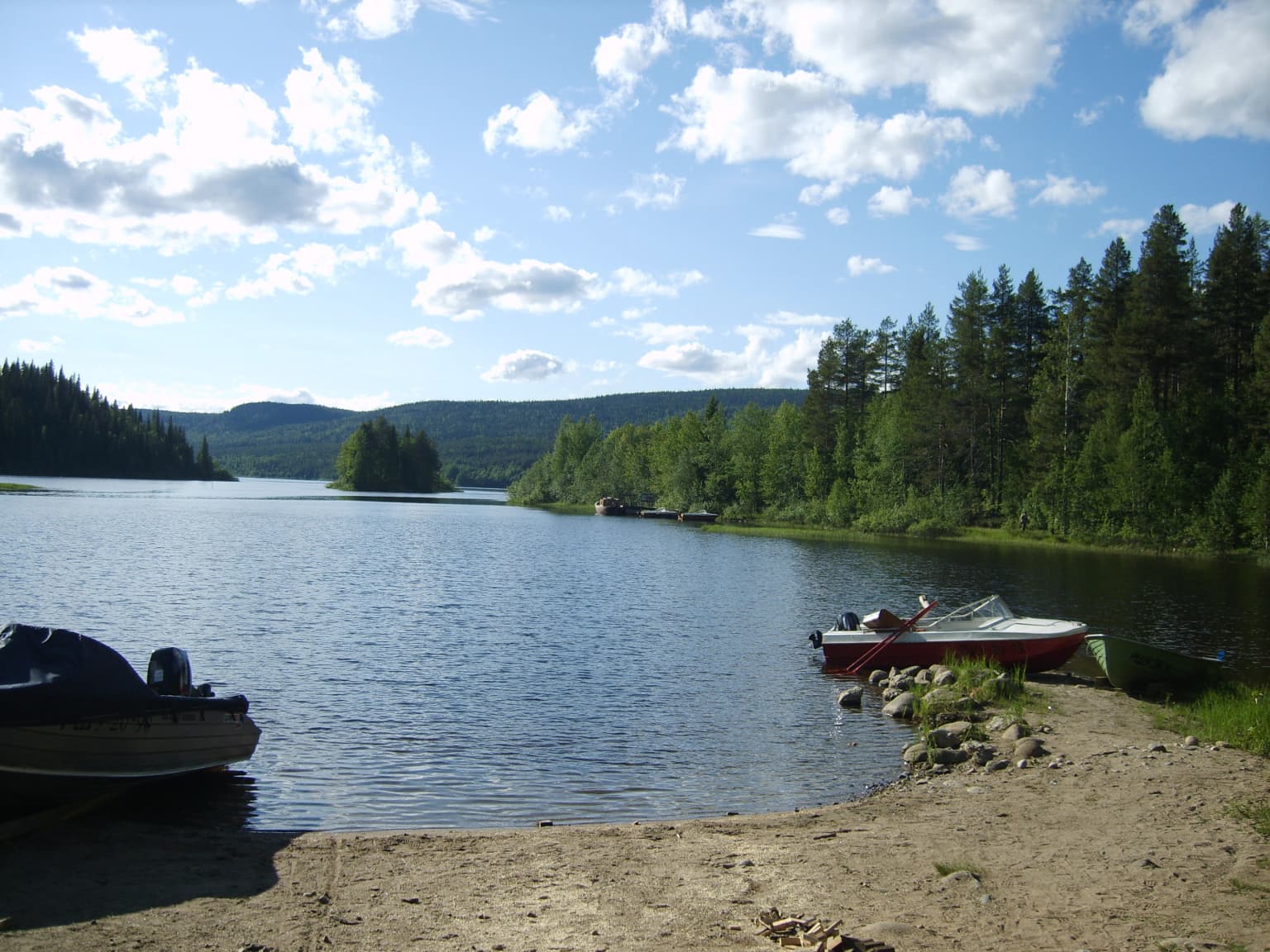 Calm lake with two motorboats on sandy shore, dense coniferous forest, distant mountains, and partly cloudy sky
