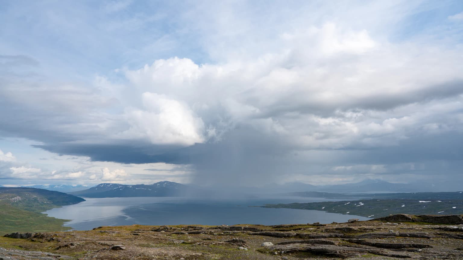 A panoramic view of a large lake surrounded by mountainous terrain under a partly cloudy sky with rain visible in the distance