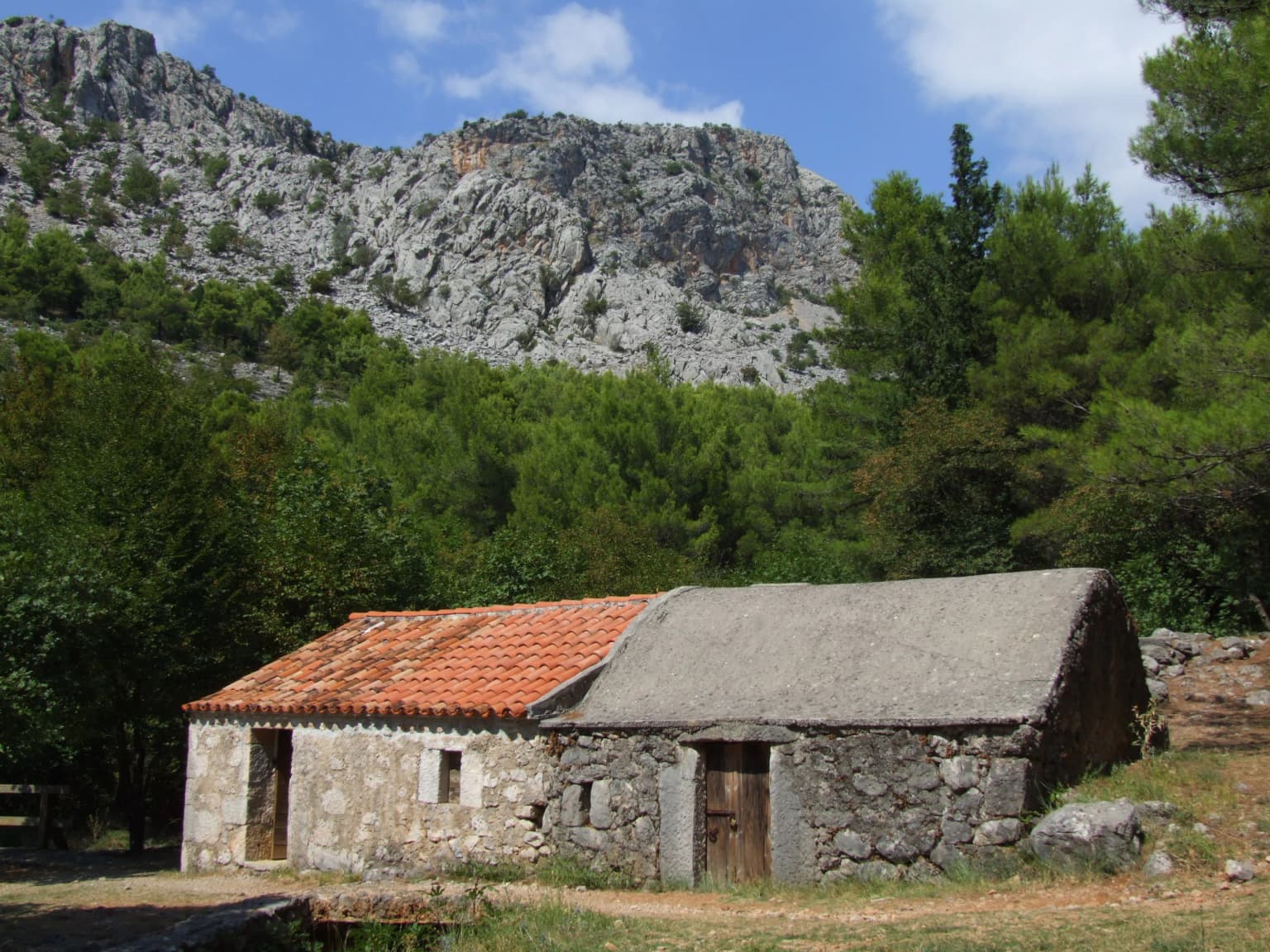 Stone building with red-tiled roof and adjacent stone structure with gray roof, set against rocky mountains and green trees