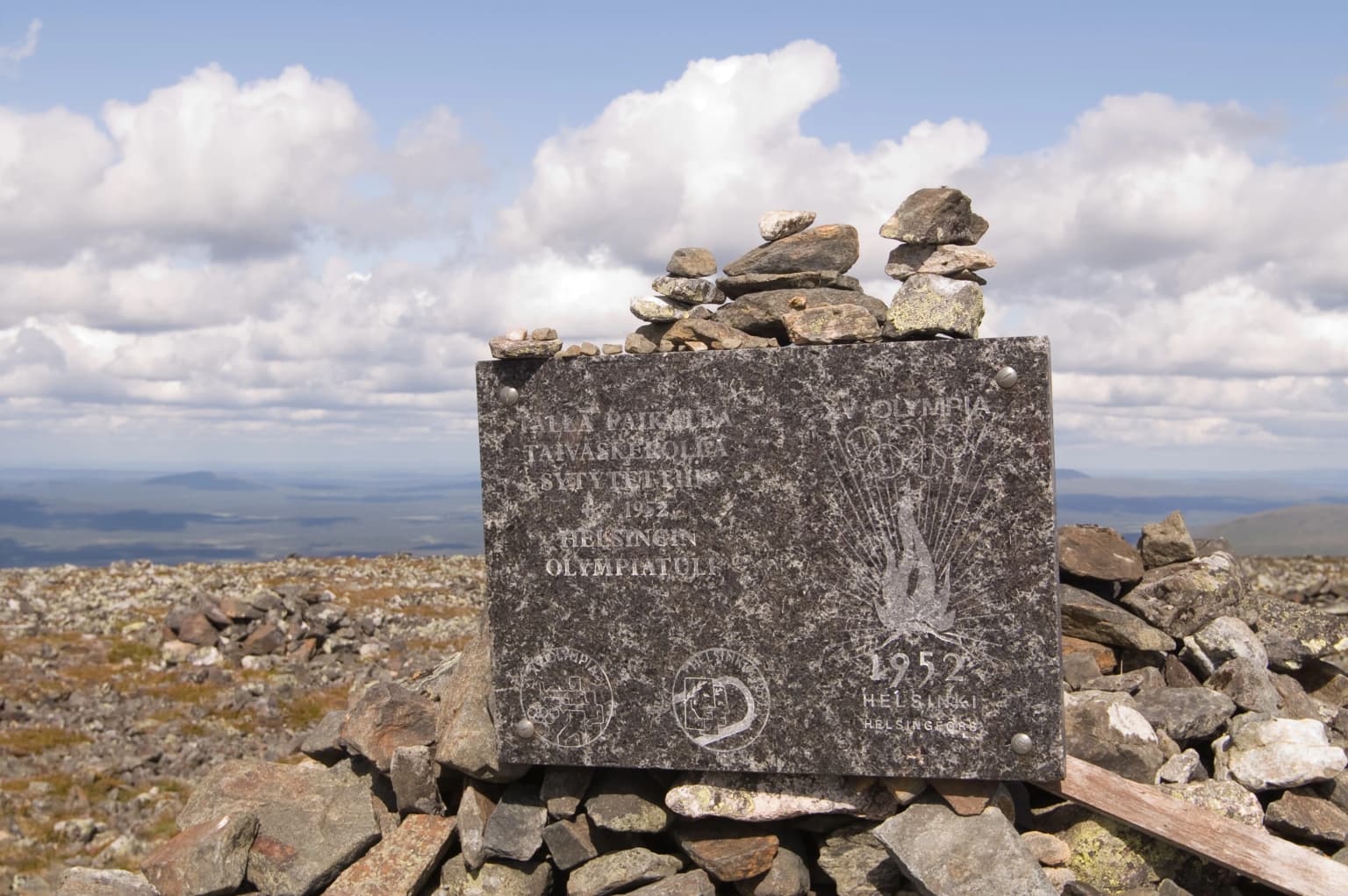 Stone monument with engraved text and Olympic flame symbol, situated on a rocky hilltop with a cloudy sky and distant landscape background