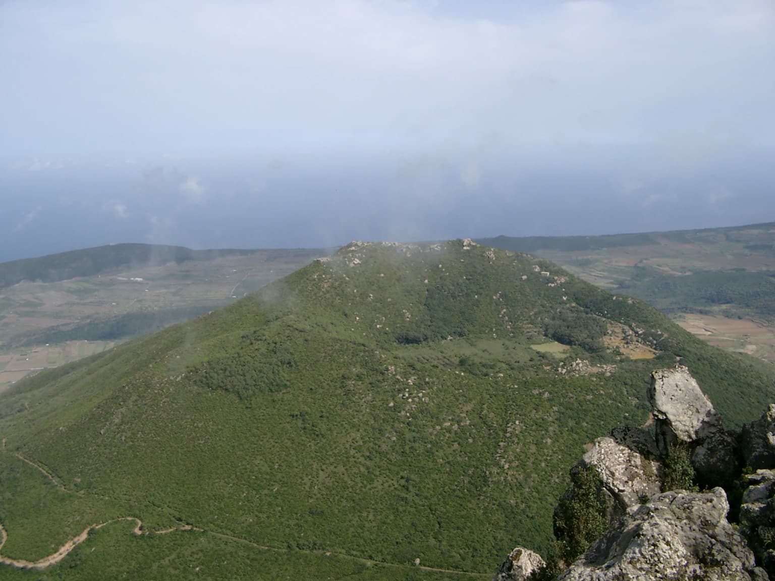 Panoramic view of a green mountain with rocky outcrops, distant hills, and a winding path under a partly cloudy sky