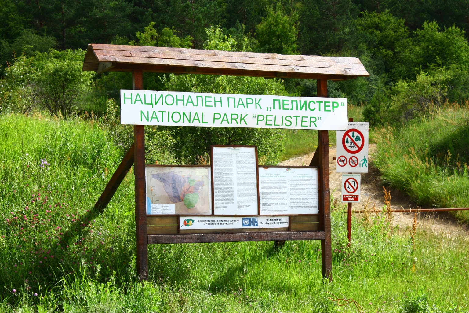 Bilingual entrance sign for National Park 'Pelister' in Macedonian and English with park information panels, surrounded by green grass and trees