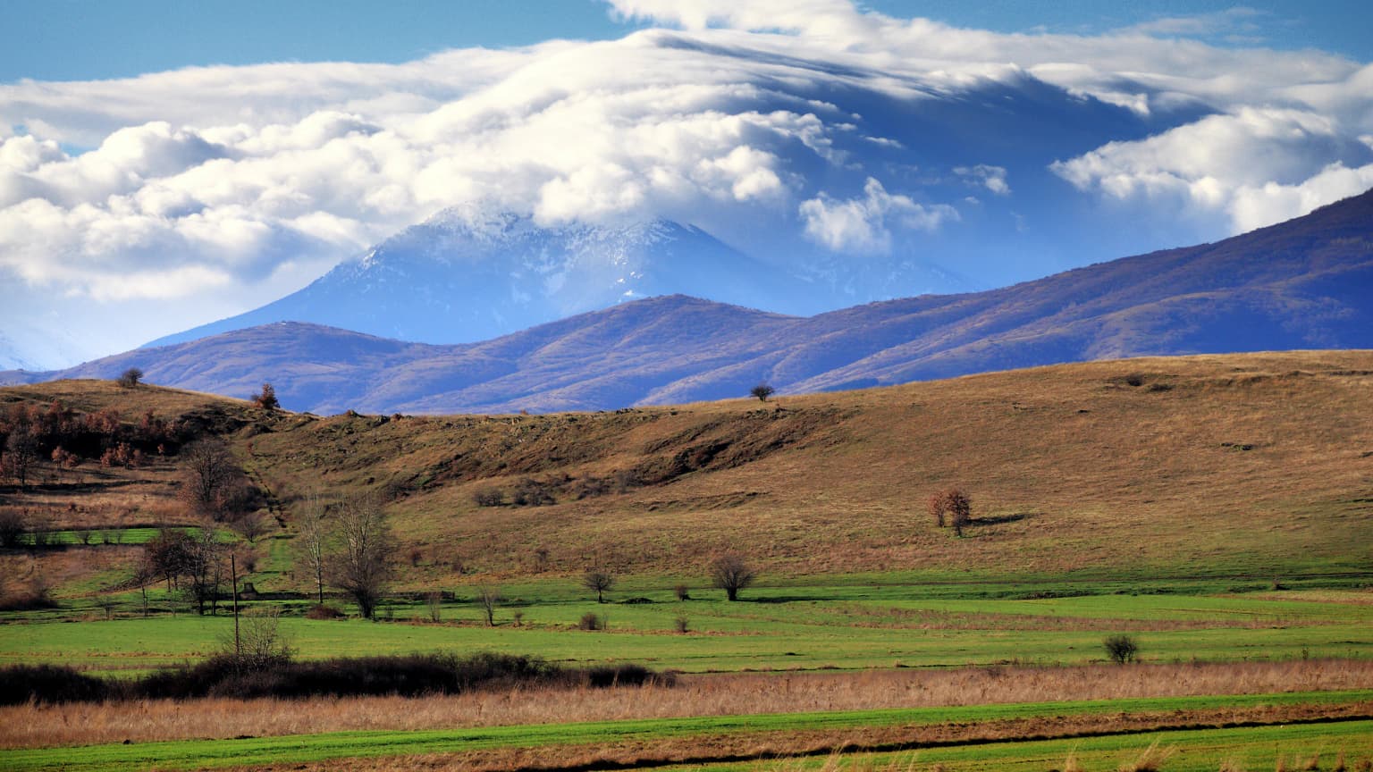 Pelister Mountain in the background with brown and green fields, scattered trees, and a partly cloudy sky
