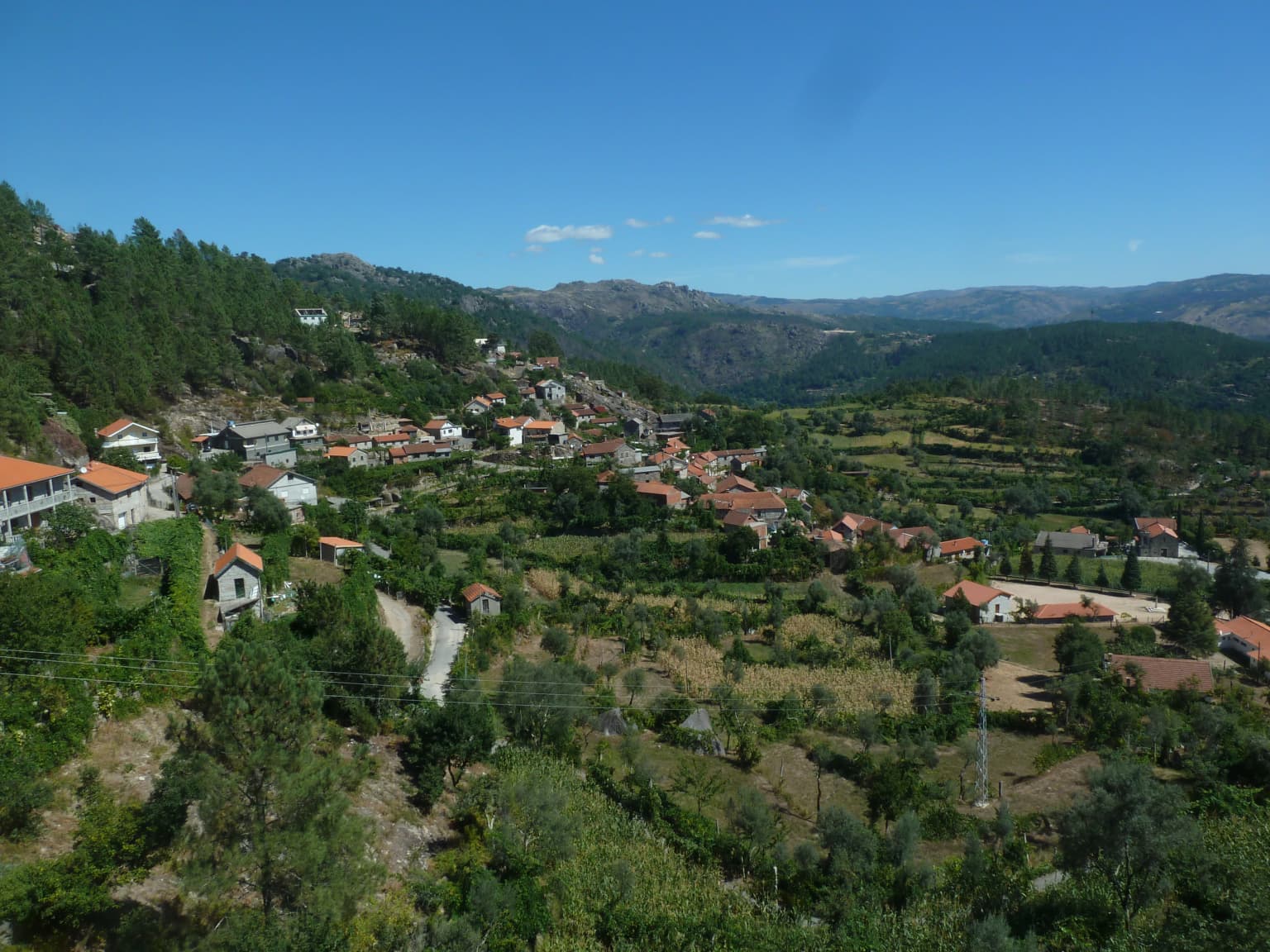 Panoramic view of a rural village with red-roofed houses, green fields, and forested hills under a clear blue sky