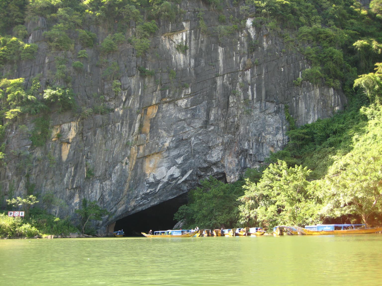 Cara de acantilado rocoso con una gran entrada a una cueva, río verde y barcos con personas en primer plano