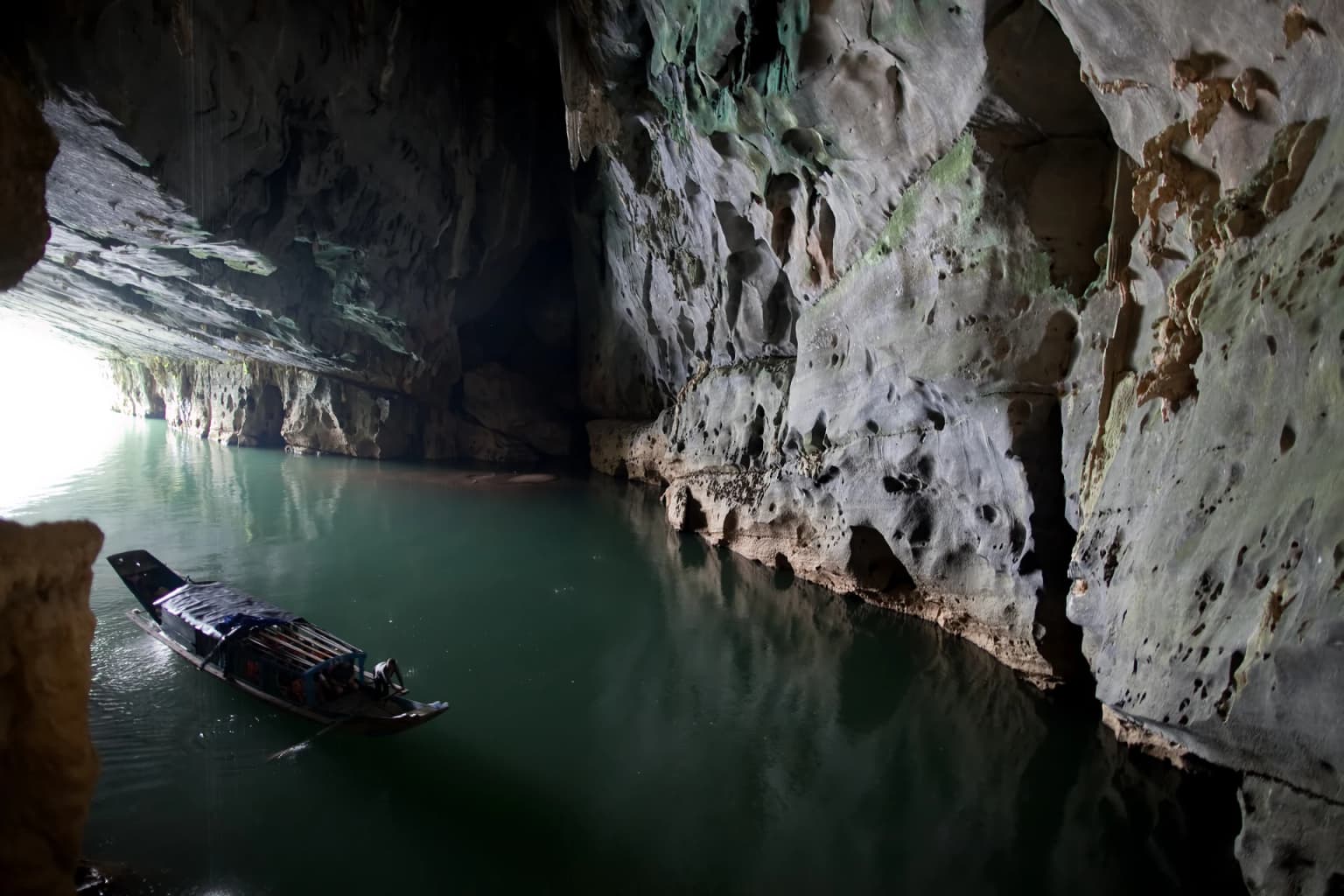 Cave interior with rocky walls, calm water, and a wooden boat entering a light-filled cave entrance