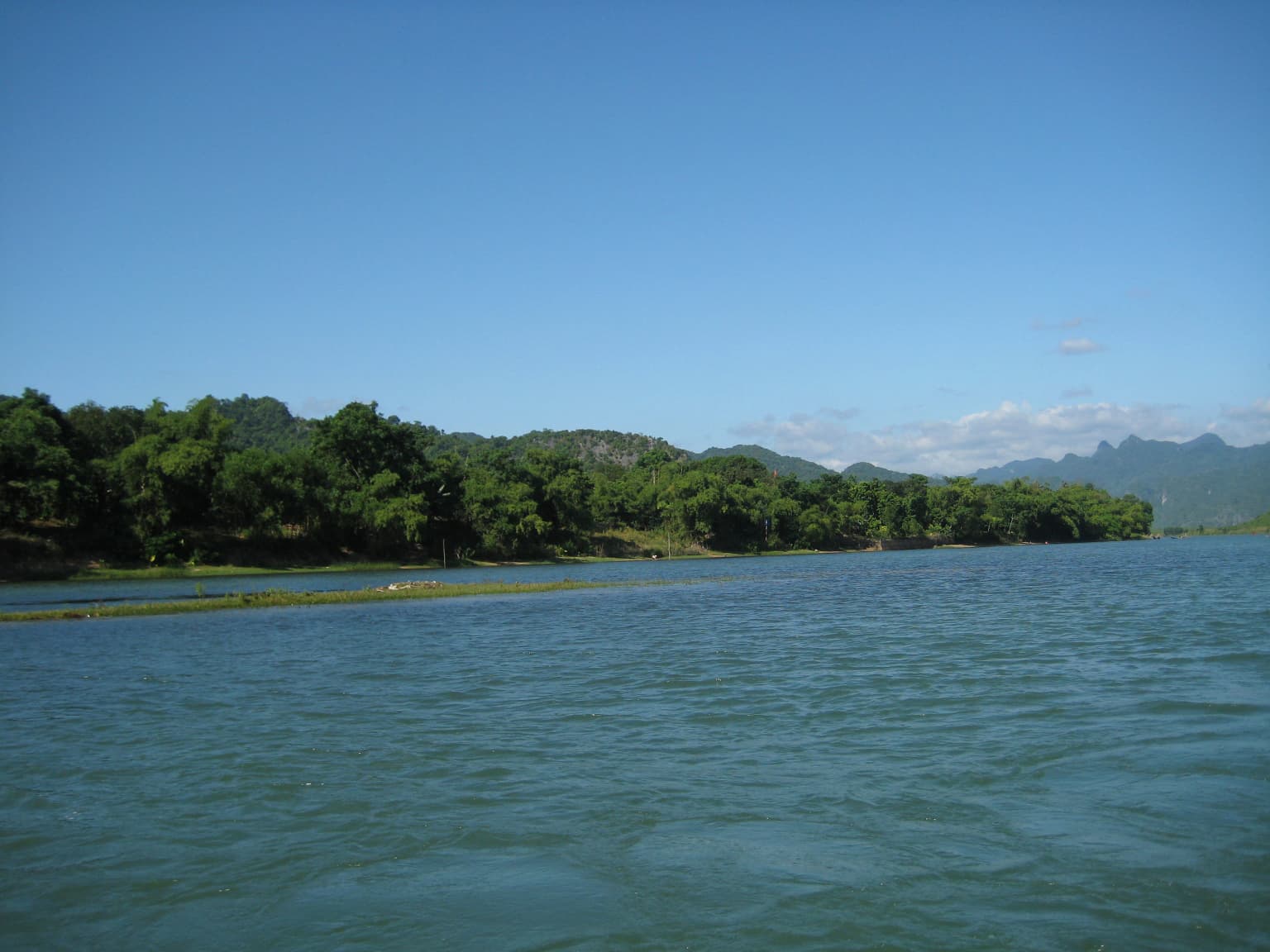 Wide shot of a river with forested shoreline and hills in the background under a clear blue sky