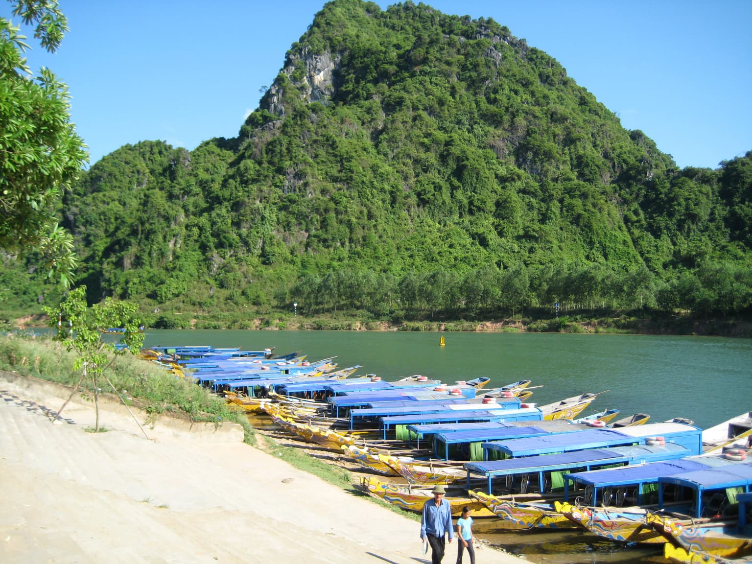 Blue-roofed tourist boats docked along a sandy riverbank with a large forested mountain in the background under a clear blue sky.